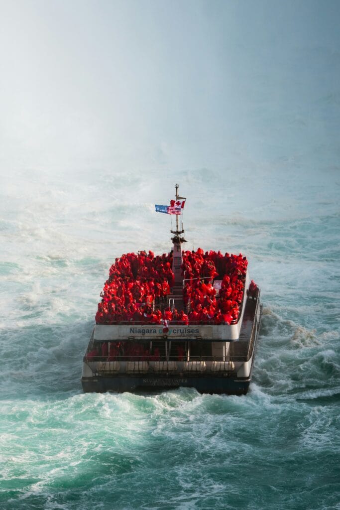 A packed Niagara Cruises boat heads toward Niagara Falls mist, its deck crowded with tourists in red ponchos
