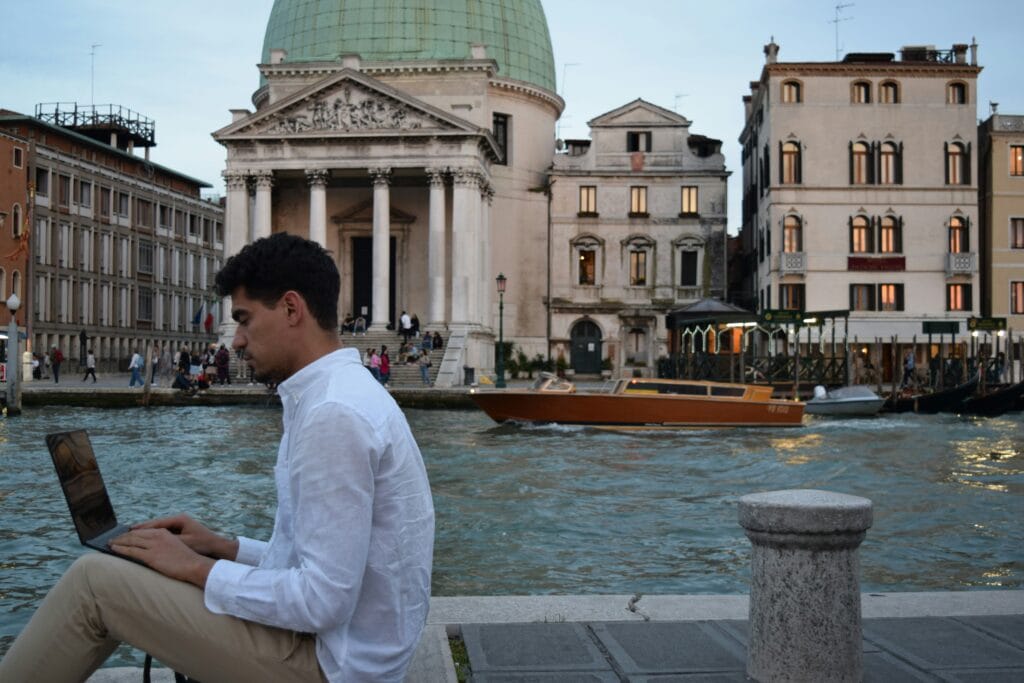 A digital nomad working on a laptop along Venice's Grand Canal, with a classic domed church and water taxi in the background