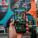 A hand holds a smartphone photographing a colorful street mural of a cat face painted on a building exterior