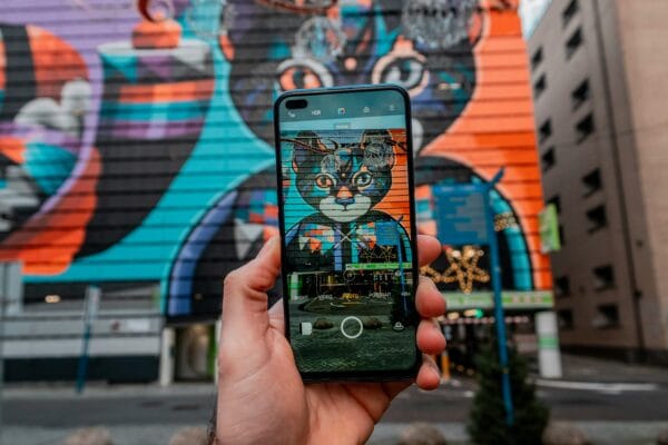 A hand holds a smartphone photographing a colorful street mural of a cat face painted on a building exterior