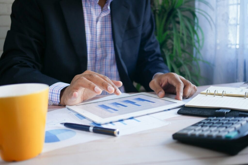 Person in a suit reviewing bar charts on a tablet at a desk with financial documents, a calculator, and a notepad nearby