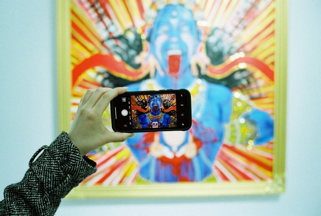 A hand holds a smartphone to photograph a vivid, colorful artwork on a gallery wall, illustrating how phones capture images that AI tools like Gemini can later search or reimagine