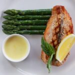 Grilled salmon fillet with steamed asparagus, a lemon wedge, fresh basil, and a small ramekin of butter sauce on a white plate