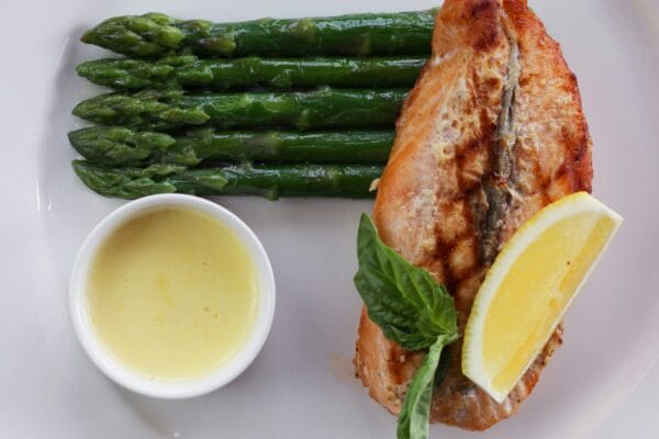 Grilled salmon fillet with steamed asparagus, a lemon wedge, fresh basil, and a small ramekin of butter sauce on a white plate