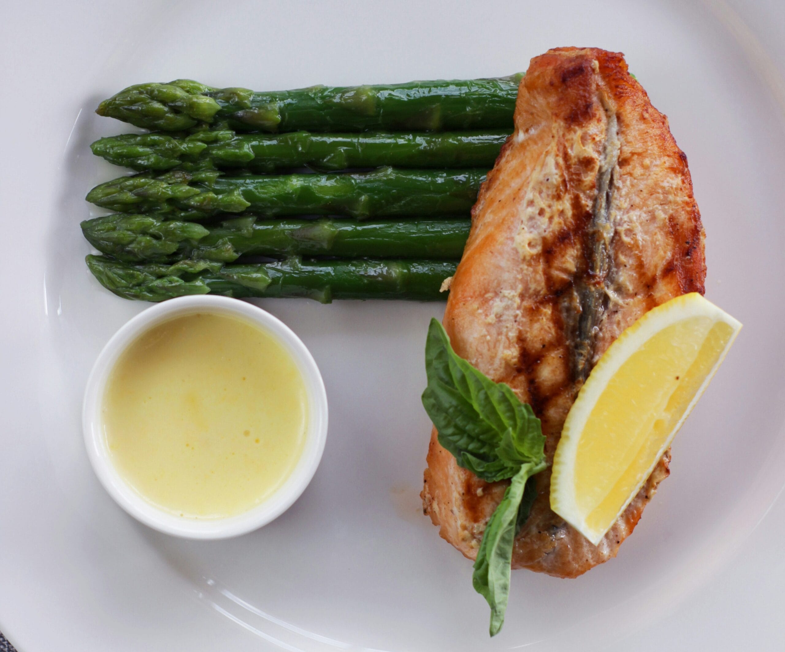Grilled salmon fillet with steamed asparagus, a lemon wedge, fresh basil, and a small ramekin of butter sauce on a white plate