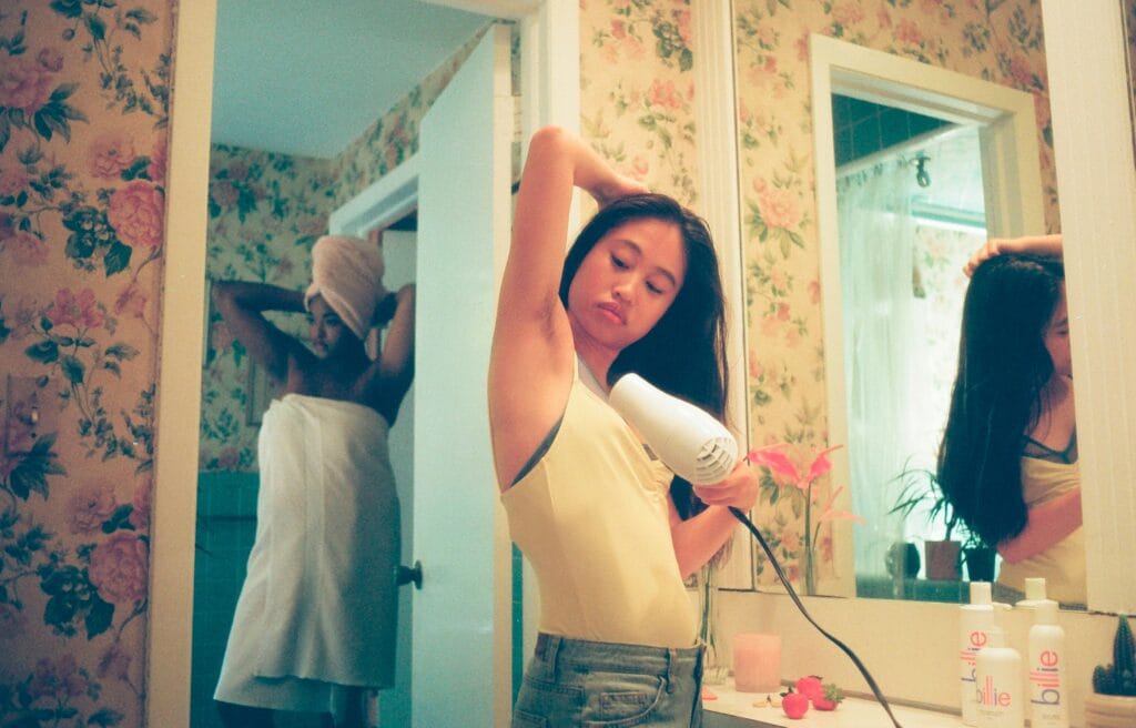 Woman blow-drying her hair in a floral-wallpapered bathroom, part of a post-shower skin and hair care routine