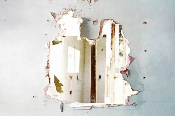 Large hole broken through a damaged interior drywall partition, exposing the room beyond. The wall shows torn plaster and jagged edges, illustrating demolition work, structural damage, or a home renovation project in progress.