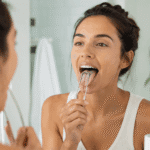 Woman using a metal tongue scraper in front of a bathroom mirror