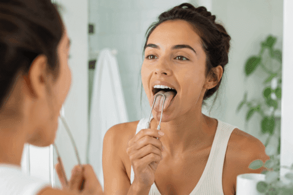 Woman using a metal tongue scraper in front of a bathroom mirror