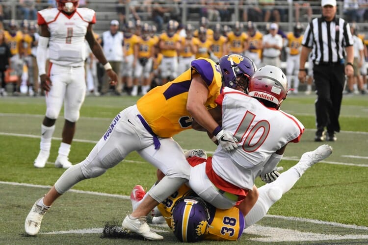 Two college football players in gold and purple uniforms tackle a ball carrier wearing number 10 in white and red during a game