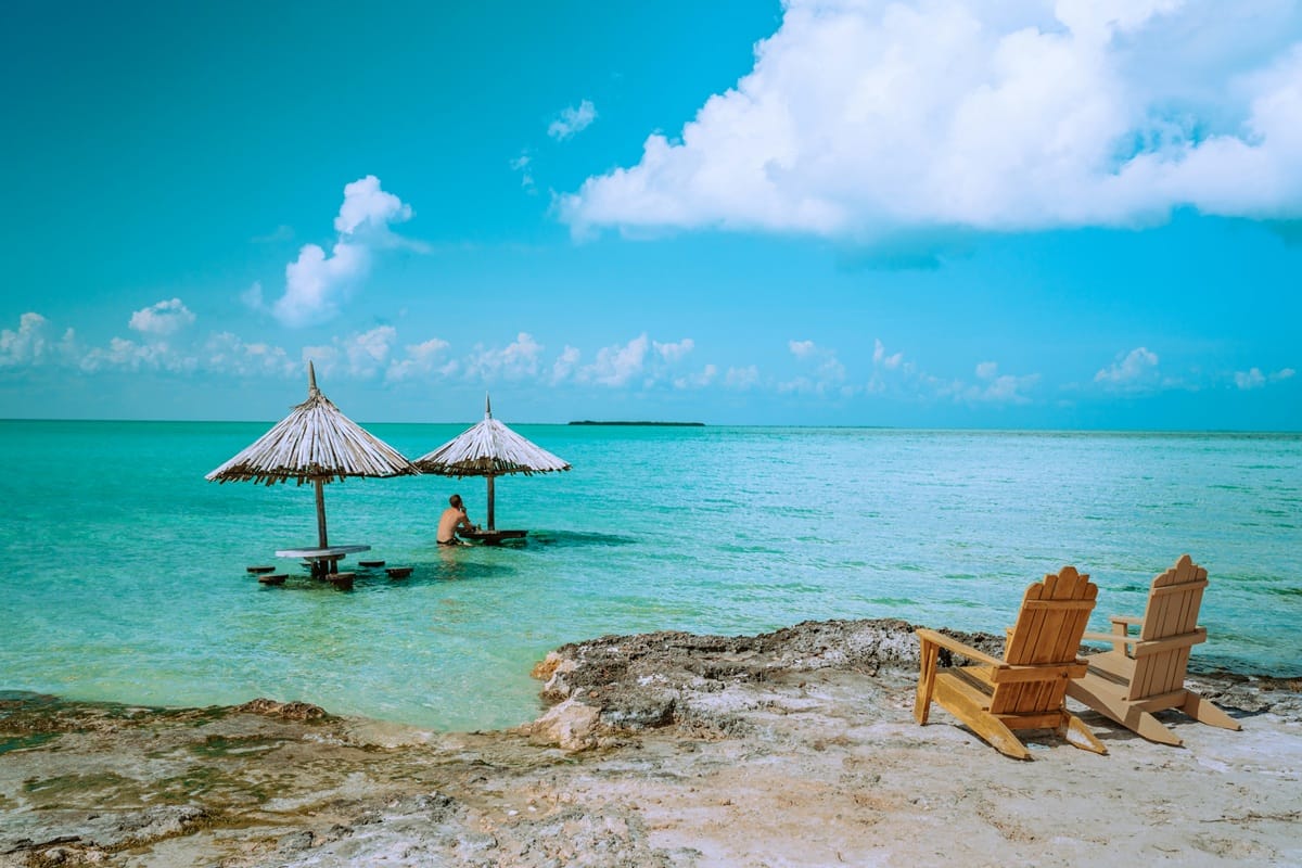 Two wooden Adirondack chairs on a rocky shore beside turquoise water, with thatched umbrella tables extending into the sea and a person sitting at one