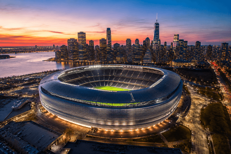 Aerial view of MetLife Stadium illuminated at dusk, with the New York City skyline visible across the Hudson River in the background