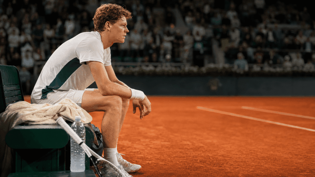 Jannik Sinner seated courtside on a clay court during a changeover, looking focused before a packed crowd