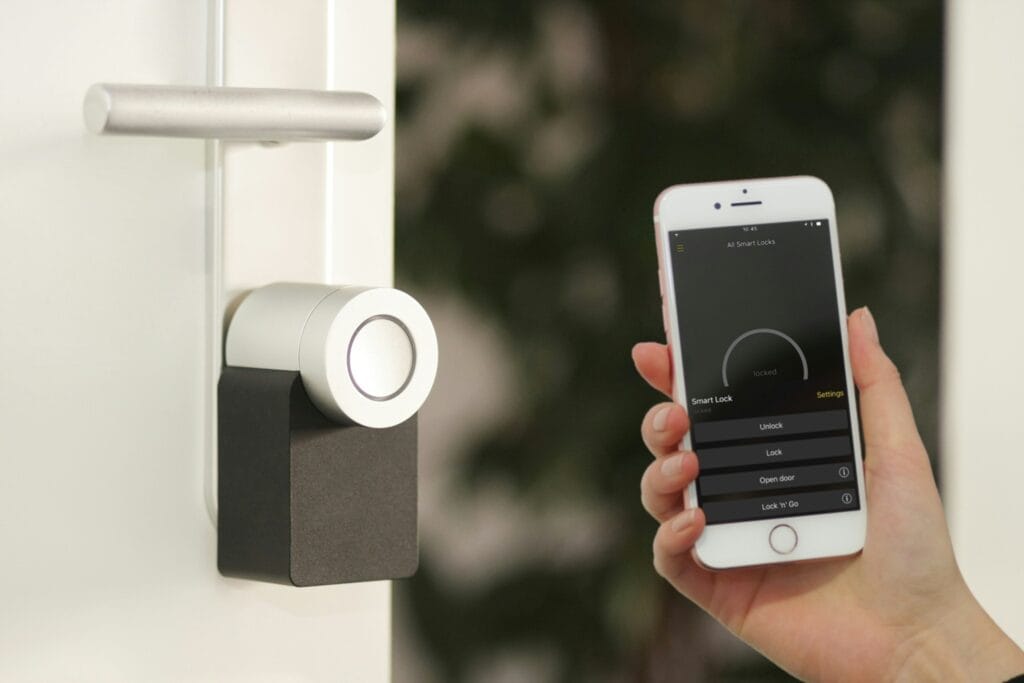 A hand holding a smartphone with a smart lock app showing lock and unlock controls, next to a smart lock mounted on a door