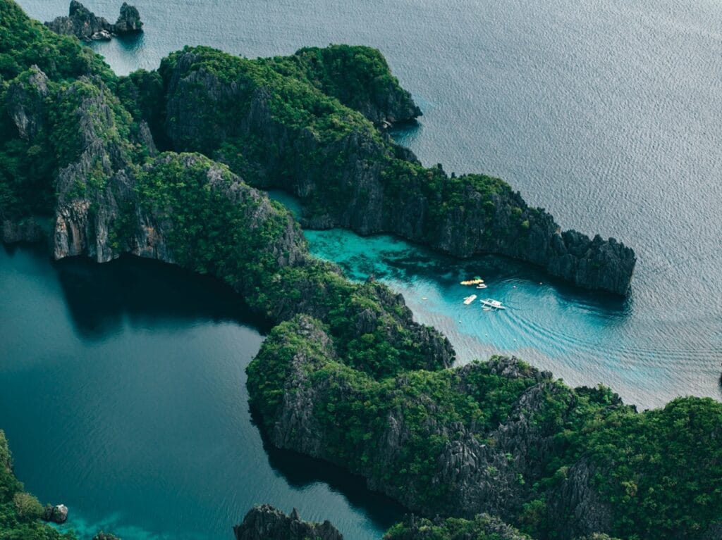 Aerial view of jagged limestone islands covered in lush greenery, enclosing a turquoise lagoon with small boats in Southeast Asia