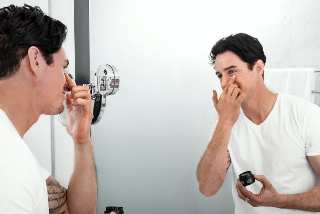 Man applying skincare product from a small jar to his face while looking in a bathroom mirror